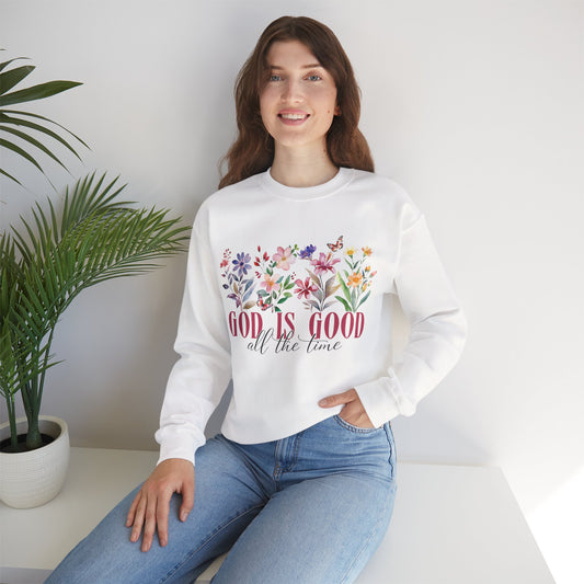 Woman wearing a white sweatshirt with floral design and 'God is good all the time' text, sitting in a room with a plant.