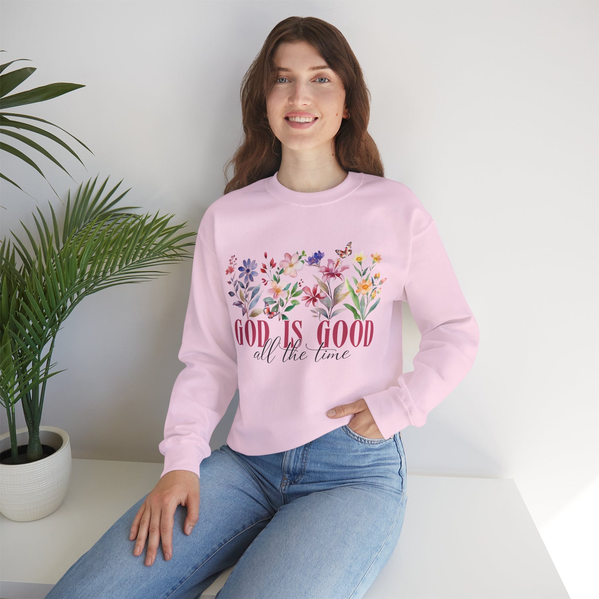 Woman wearing a pink sweatshirt with floral design and text, sitting on a white surface with a plant in the background.