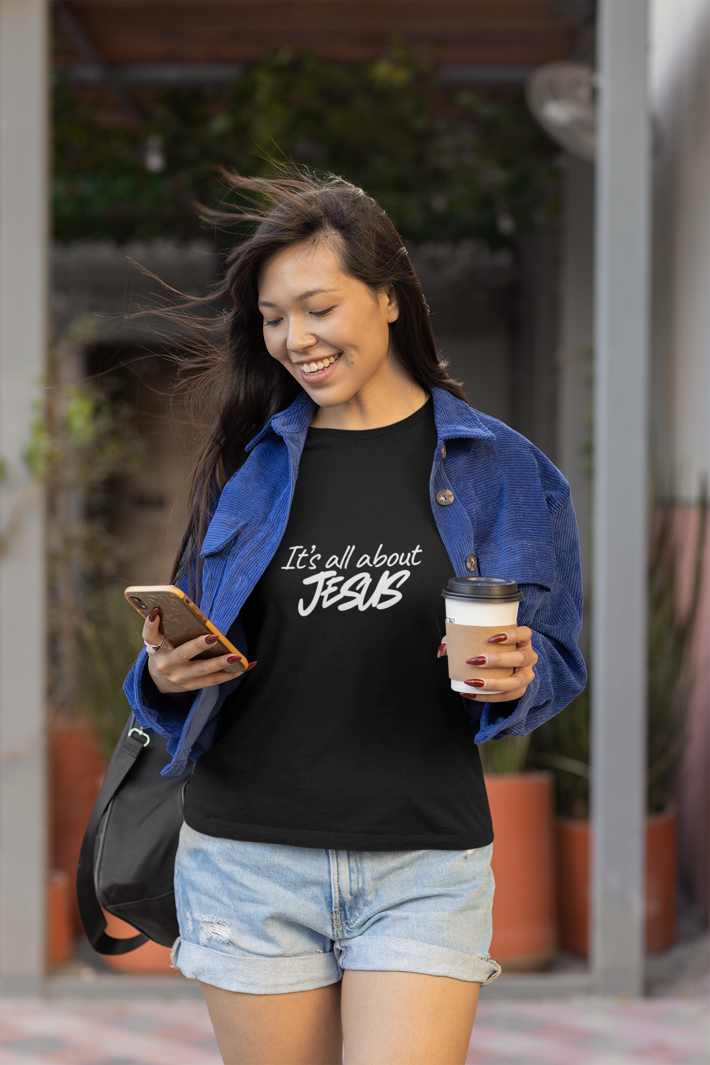 Woman walking outdoors, holding a phone and a drink, wearing a black t-shirt with text.