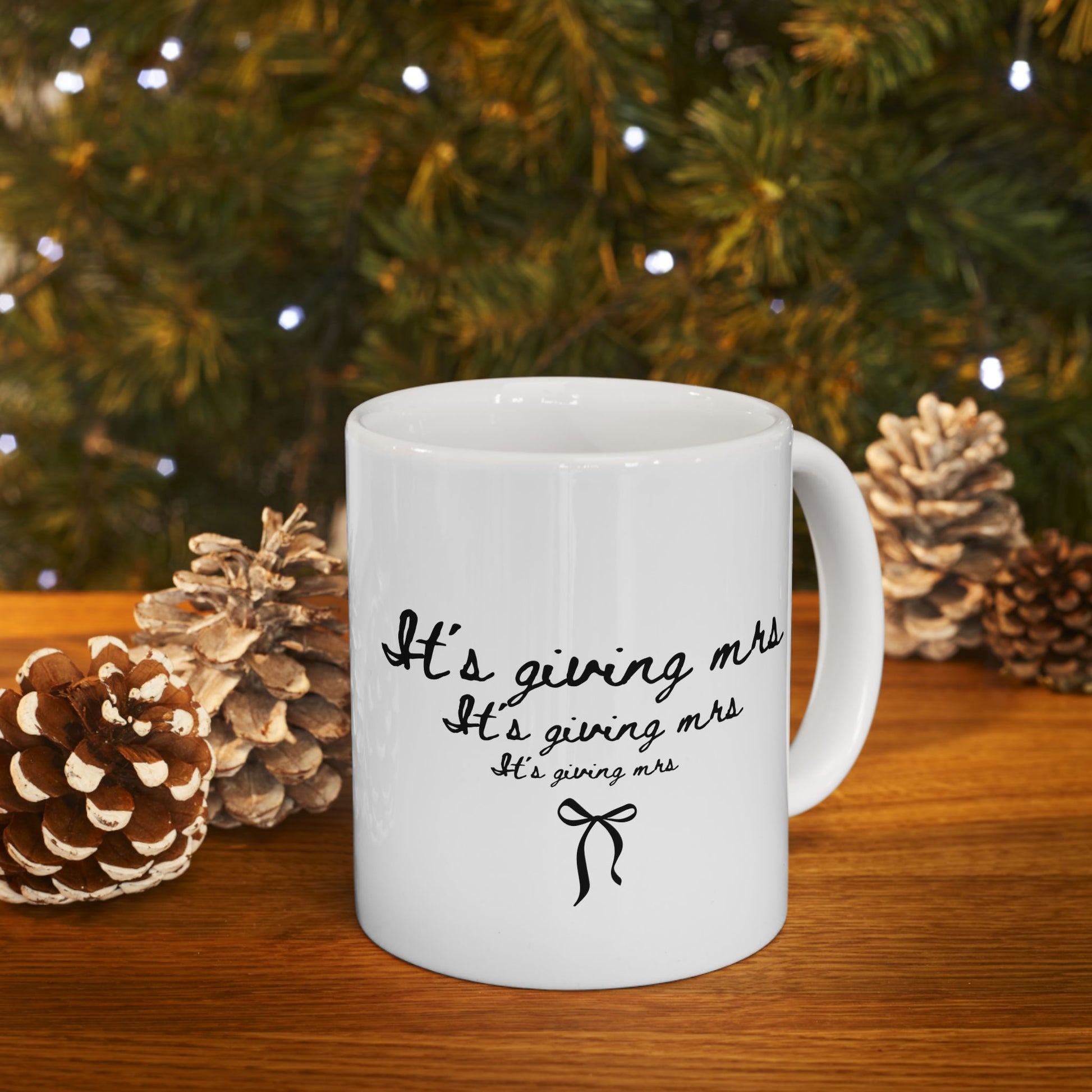 White mug with text in front of a Christmas tree and pinecones